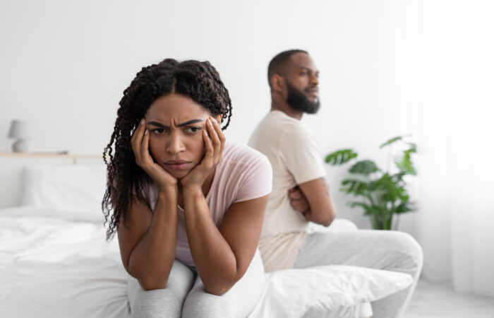 Upset millennial african american wife ignores offended husband on white bed in minimalist bedroom